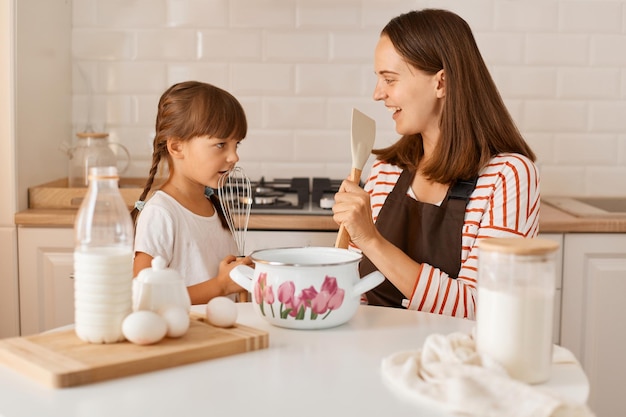 Extremely happy excited young adult woman cooking with her daughter at home, having fun together, singing with kitchen equipment, enjoying spending time together.