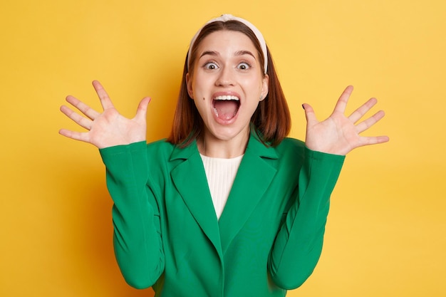 Extremely happy surprised woman wearing green jacket posing isolated over yellow background raised her arms screaming with happiness excited emotions