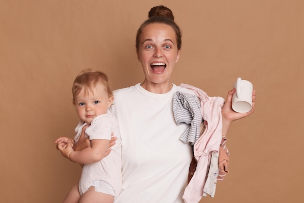 Extremely happy young mother with bun hairstyle holding her toddler daughter and screaming happily rejoicing hearing good news posing isolated over brown background