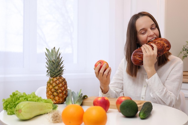 Extremely hungry brown haired Caucasian woman wearing white shirt sitting on table in light kitchen interior holding apple biting sweet baked pie breaking diet