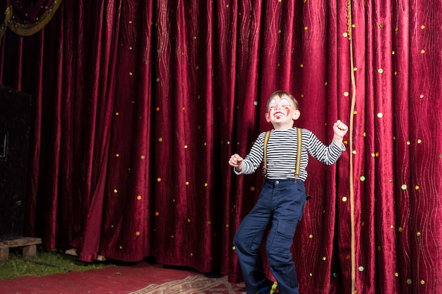 Exuberant little boy performing on stage at a pantomime standing in front of the closed burgundy curtains doing his act in his costume and makeup with a laughing smile, with copyspace