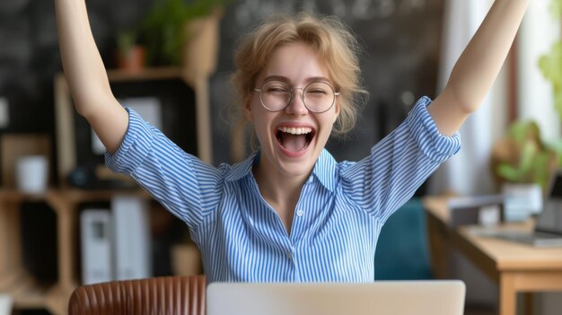 exuberant woman with tousled hair and glasses is raising her arms triumphantly in front of a laptop expressing joy and success