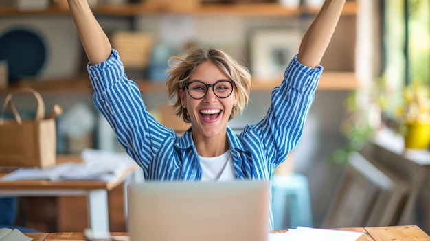 exuberant woman with tousled hair and glasses is raising her arms triumphantly in front of a laptop expressing joy and success