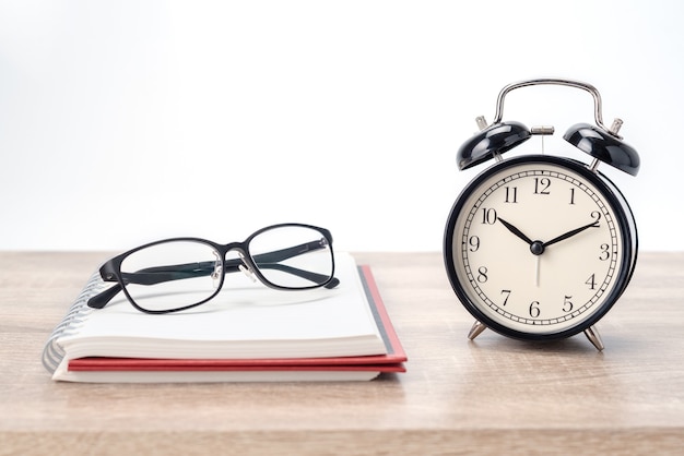 eyeglasses on a notebook and an alarm clock on wooden table and white background