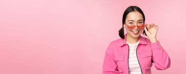 Eyewear advertisement Stylish modern asian girl touches sunglasses wears pink poses against studio background Copy space