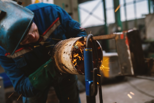 Photo fabric worker in protective uniform cutting metal pipe on the work table with an electric grinder in the industrial workshop.