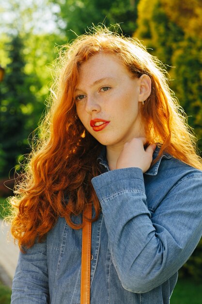 Fabulous redhead young model in blue denim shirt posing  in rays of sun at the park