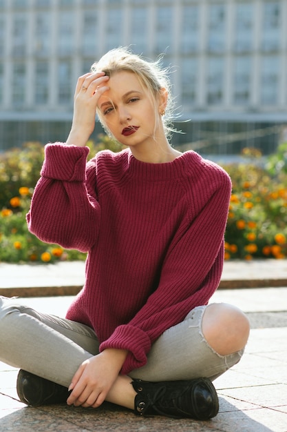 Fabulous young model wearing knitted sweater and torn jeans sitting on the sidewalk