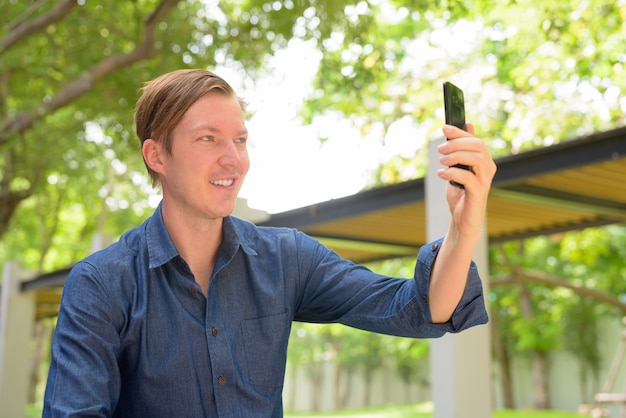 Face of happy young handsome blond businessman taking selfie at the park outdoors