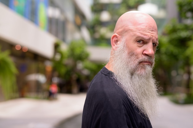 Face of mature bald bearded man looking serious at camera with nature in the city