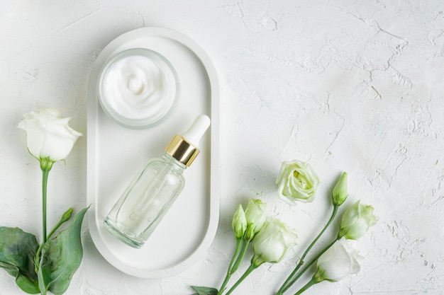 Face moisturizer cream serum or essential oil in glass bottles and eustoma flowers on white concrete background Flat lay top view Set for skin and body care beauty products