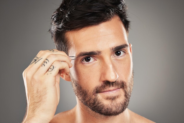 Face portrait and tweezers with a man model plucking his eyebrows in studio on a gray background Skincare wellness and luxury with a handsome young male tweezing his eyebrow hair for grooming