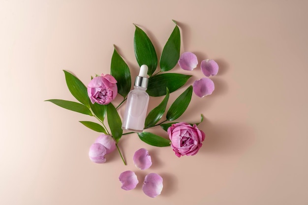 A face serum or essential oil in a pink dropper bottle lying on a beige background with rose petals and green leaves around it