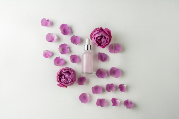 A face serum or essential oil in a pink dropper bottle lying on a white background with rose petals around it