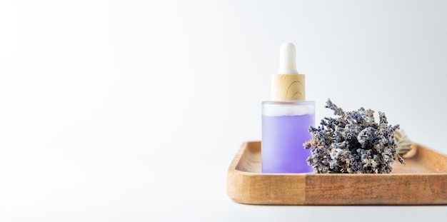 A face serum or oil in a transparent dropper bottle on a wooden tray on white background with lavender flowers nearby Banner