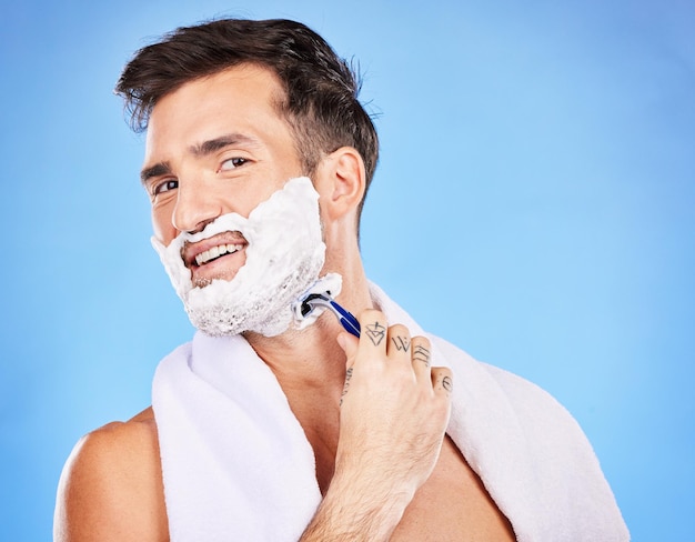 Face shave and hair removal with a man model shaving his skin in studio on a blue background for wellness or hygiene Portrait beard and razor with a young male grooming alone in the bathroom