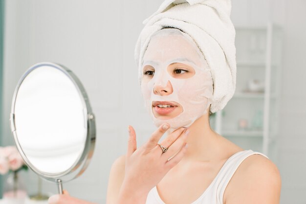 Face skin care and beauty treatments. Woman with a sheet moisturizing mask on her face and white towel on the head, looking at the mirror.