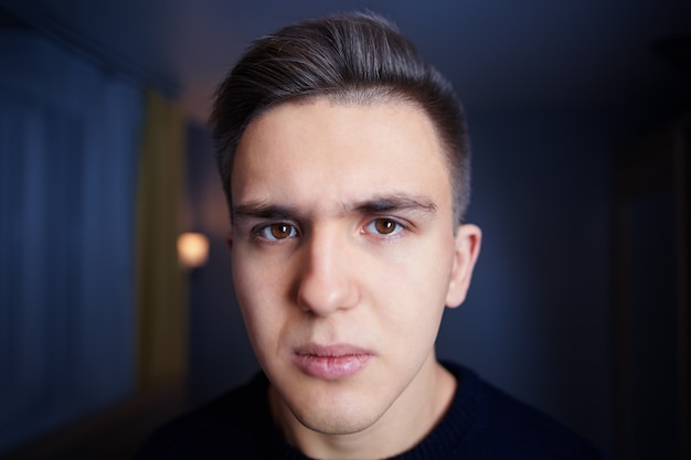 Face of young white man with brown eyes, short hair, and sullen look, against the wall of dark blue room.