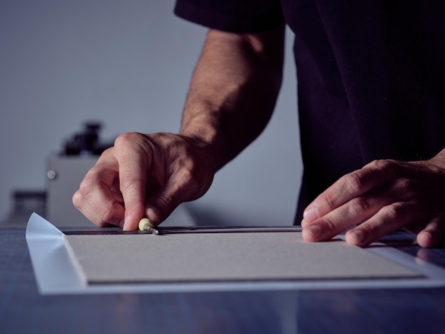 Faceless craftsman pressing adhesive layer to cardboard base with special tool while making book bind