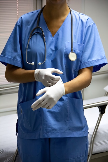 Faceless portrait of a doctor in a light blue uniform putting on sterile gloves, holding a stethoscope.