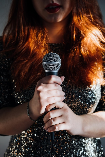 Faceless portrait of redhead woman in sparkly evening dress holding microphone on dark night