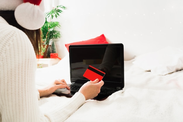 Faceless woman in santa hat lying on bed at home with laptop and plastic card