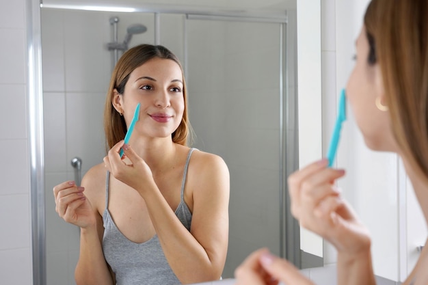 Facial hair removal Beautiful young woman shaving her face by razor at home Pretty woman using shaver on bathroom