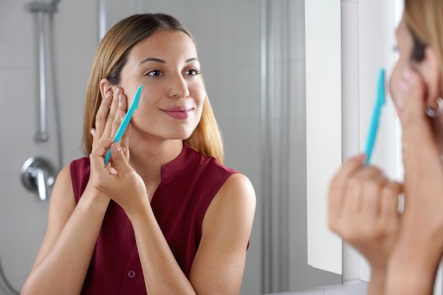 Facial hair removal Brazilian girl shaving her face by razor at home Beautiful young woman using razor on bathroom