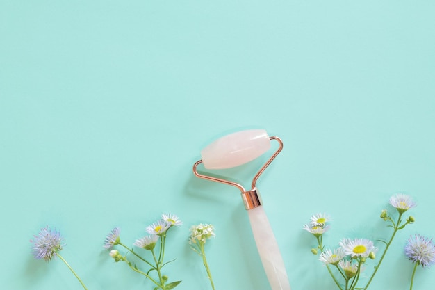 Facial massage roller made of rose quartz on a blue background with flowers Flat lay Spa