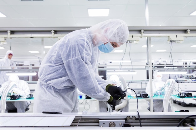 A factory worker in a white lab suit black latex gloves and face mask working with a soldering iron other workers are on the background