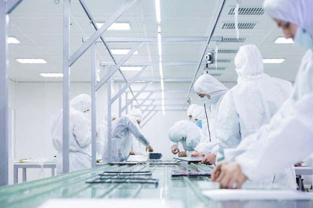 Factory workers in white lab suits and face masks producing tv sets on a green assembly line with some modern equipment