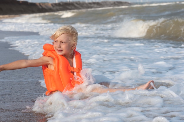 Fair-haired boy in an inflatable swimming vest sits on seashore in the water