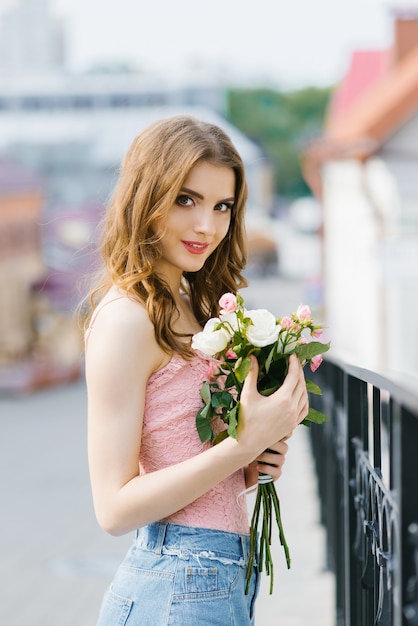 A fair-haired young beautiful pretty girl in summer clothes stands and holds delicate white and pink roses in her hands after a date