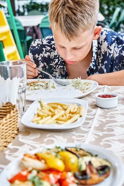 Fairhaired boy eats tasty food sitting at outdoor table with plates of dishes closeup