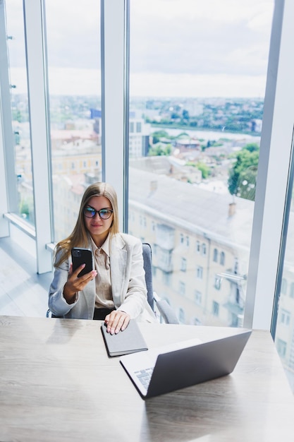 A fairhaired cheerful European woman in glasses in stylish casual clothes is sitting at a table with a laptop doing paperwork and talking on the phone Business lady at the workplace in the office