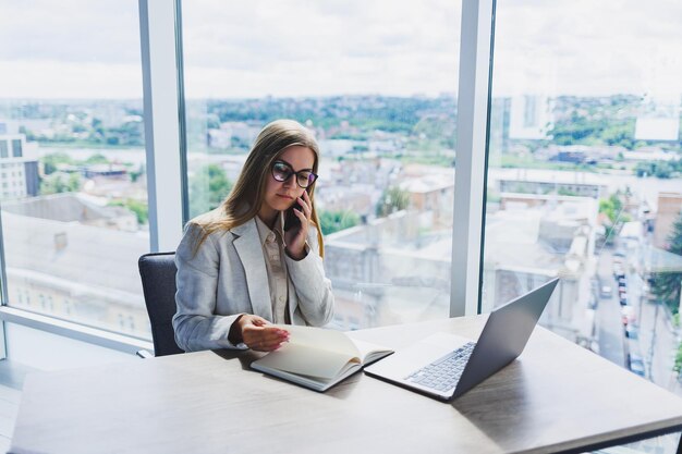 A fairhaired cheerful European woman in glasses in stylish casual clothes is sitting at a table with a laptop doing paperwork and talking on the phone Business lady at the workplace in the office