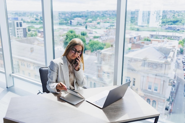 A fairhaired cheerful European woman in glasses in stylish casual clothes is sitting at a table with a laptop doing paperwork and talking on the phone Business lady at the workplace in the office