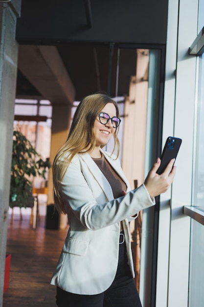 A fairhaired European woman in a white jacket and glasses is talking on the phone while standing in a spacious office with large windows Successful young woman freelancer