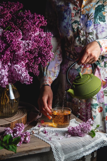 A fairskinned girl in a pastelcolored dress pours tea from a green castiron teapot into a glass Spring composition with lilacs on a vintage table Voluminous soft shadows Low key