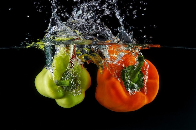 Falling Bulgarian sweet orange and green peppers into the water, spray for design, freezing in motion. Water splash and vegetables isolated