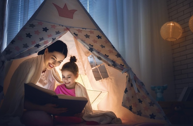 Family bedtime. Mom and child daughter are reading a book in tent. Pretty young mother and lovely girl having fun in children room.