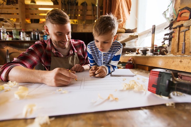 family, carpentry, woodwork and people concept - happy father and little son with blueprint at workshop