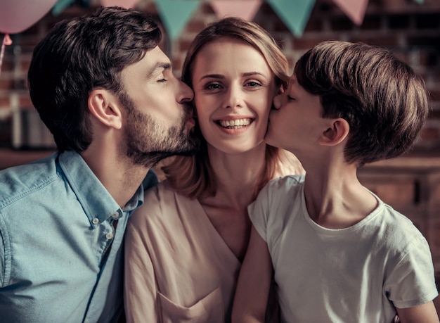 Family celebrating mother's birthday in decorated kitchen. Beautiful woman is looking at camera and smiling while husband and son are kissing her in cheeks