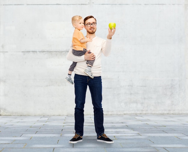 family, childhood, fatherhood, healthy eating and people concept - happy father and and little son with green apple over urban street background