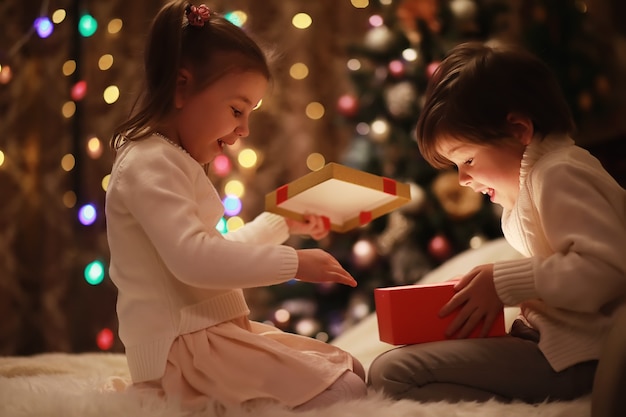 Family on Christmas eve at fireplace. Kids opening Xmas presents. Children under Christmas tree with gift boxes. Decorated living room with traditional fire place. Cozy warm winter evening at home.