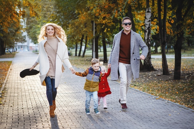 Family dad, mom, son and daughter are running along a pavement alley in a park. He is in a gray coat and black glasses, she is in white, the girl is blonde in red.