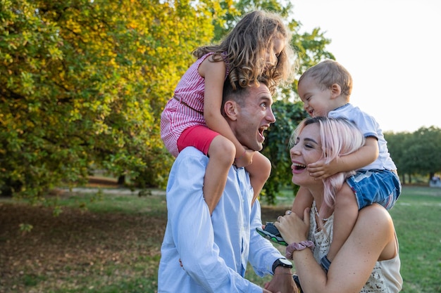 Family of four at the park Babies on shoulders dad opens wide his mouth wide and makes mom and babies laugh