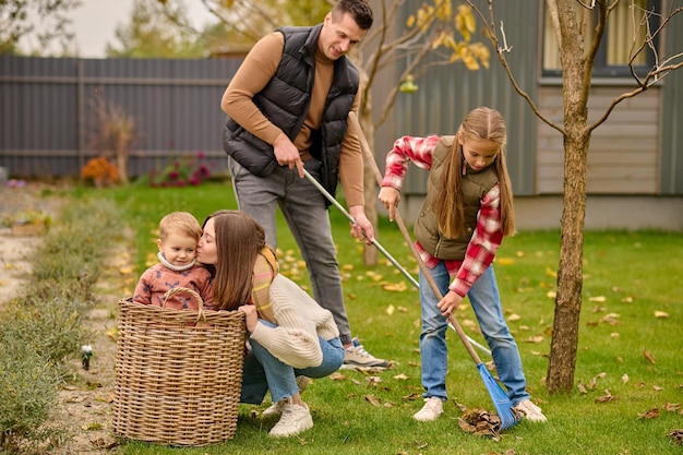 Family, garden. Smiling man with school-age girl raking leaves young blonde woman crouching kissing cute child in basket in garden on autumn day