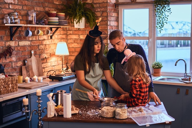 Family in halloween. Mom dad and little daughter with makeup and hats cooking together in loft style kitchen at morning.