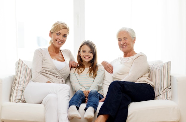 family, happiness, generation and people concept - smiling mother, daughter and grandmother sitting on couch at home